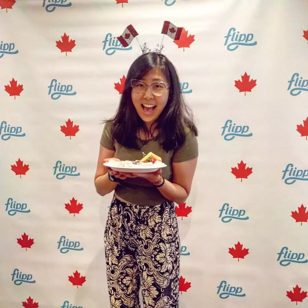 Smiling girl holding a plate while standing in front of a backdrop interspersing a Canadian maple leaf and the Flipp logo, wearing a headband prop with Canadian flag antennae