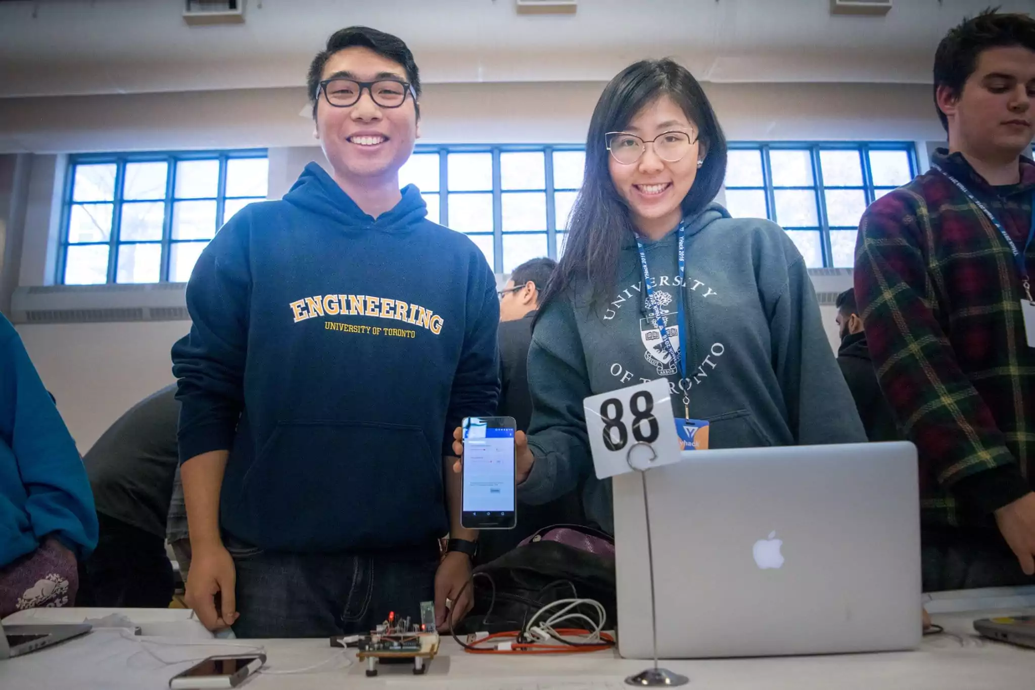 Two students at a Hackathon standing with an electronic device (Arduino, breadboard, wires), holding a phone with an app
